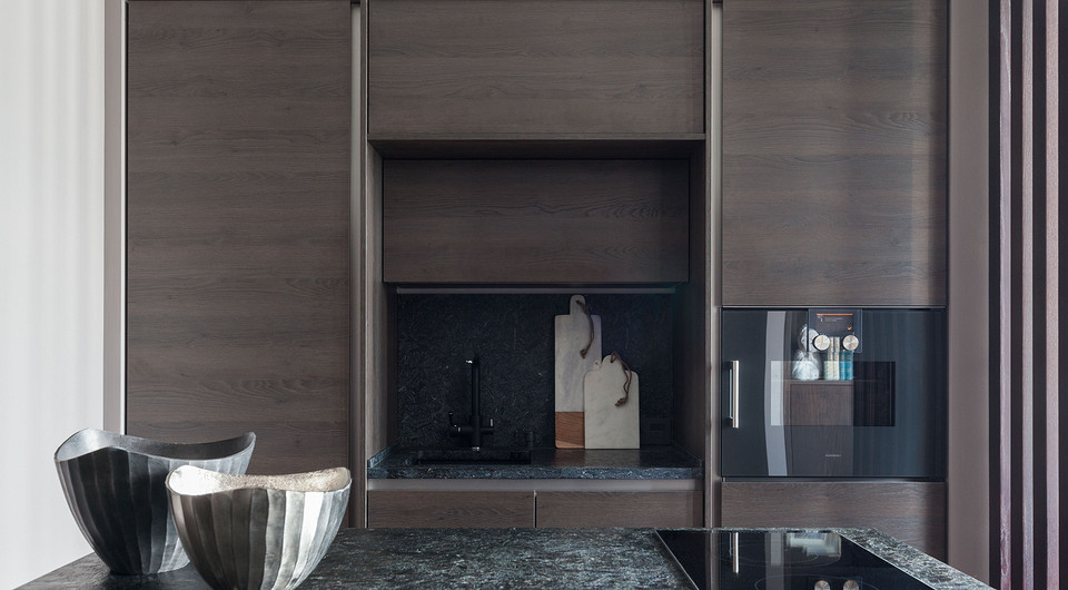 Кухонные модули встроены в нишу, которую образуют конструкции из гипсокартона. В верхней части скрыт рукав вытяжки, в боковую со стороны подиума встроен стеллаж. В нижней части подиума пр...