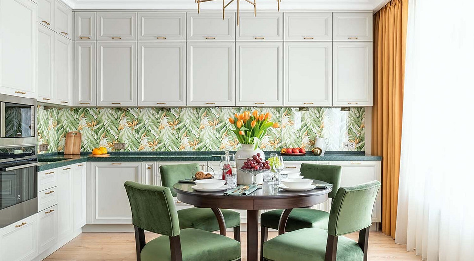 Обеденный стол стал графичной доминантой общего пространства. Зелёные стулья и кухонный фартук с буйным растительным принтом привносят ощущение тропиков, а оранжевые шторы — солнечну...