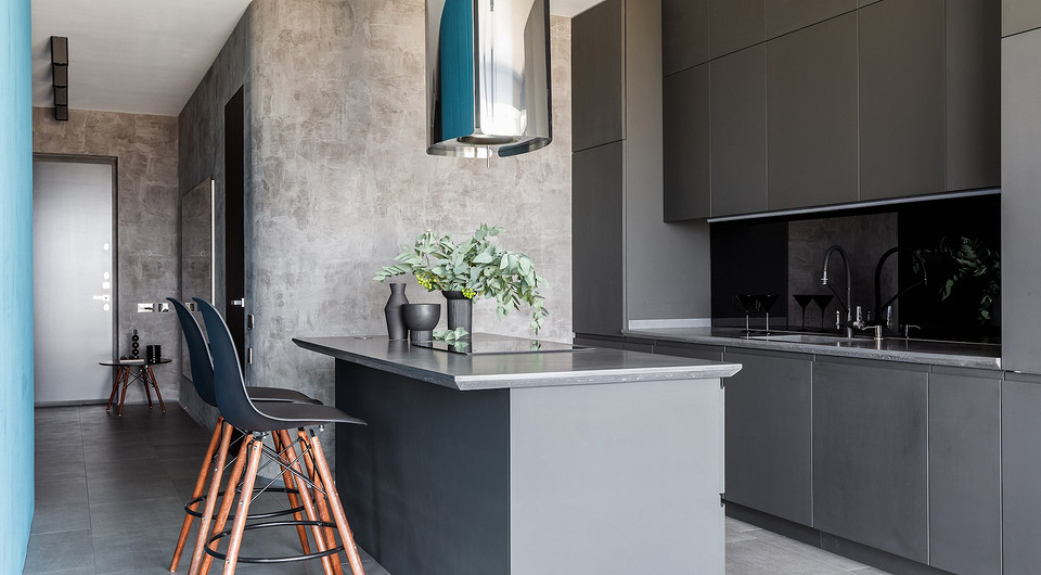 Графитовая кухня с островом выделяется на фоне светло-серых стен и объединяется по цвету с обеденным столом и потолочными светильниками.